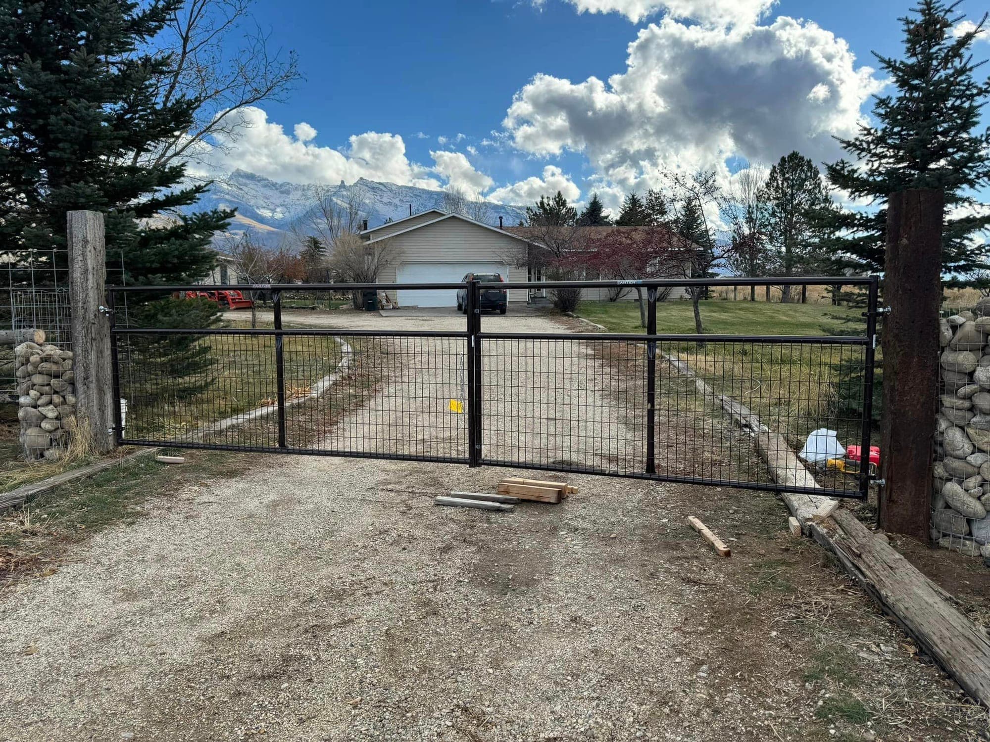 Black steel wire double driveway gate hung on rock-filled gabion pillars with the snowy Ruby Mountains behind it, Elko, Nevada