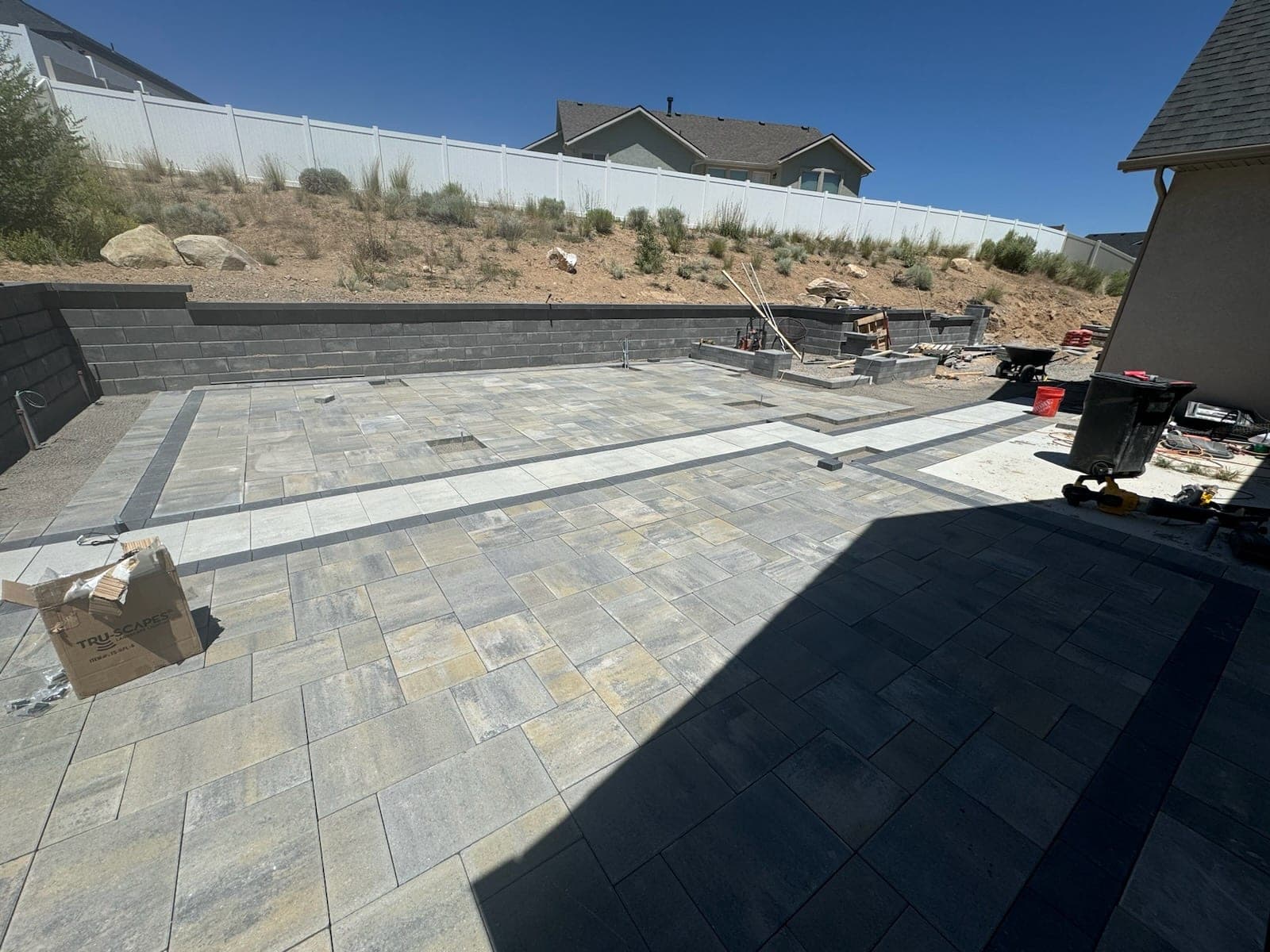 Large paver patio with a dark border being laid below a block retaining wall on a sloped lot, Elko, Nevada