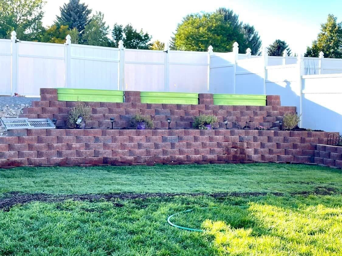 Tiered block retaining wall with raised garden beds, landscape lights and new sod in front of a white vinyl fence, Elko, Nevada