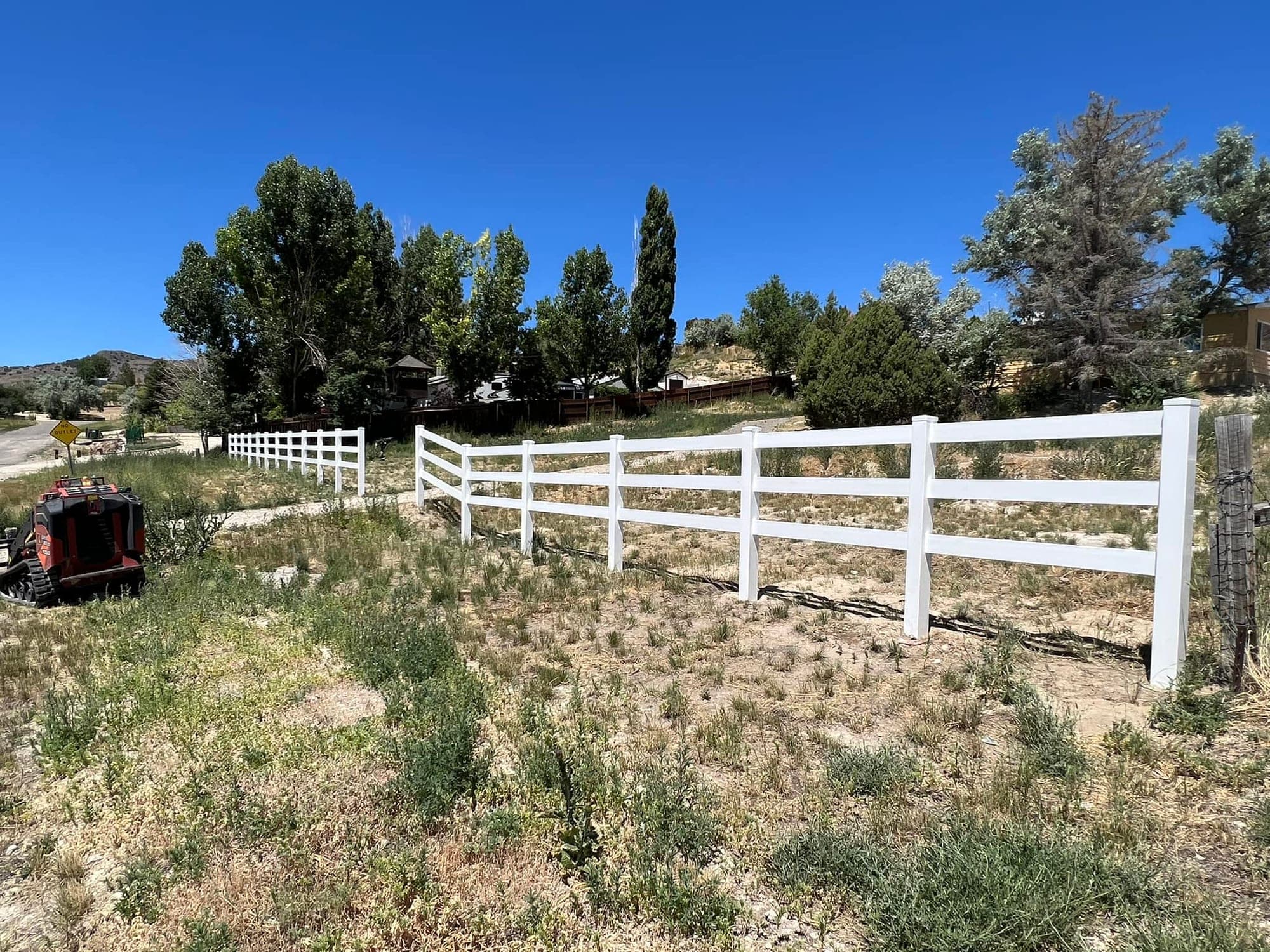 White three-rail vinyl ranch fence running along a driveway with juniper and cottonwood trees, Elko, Nevada